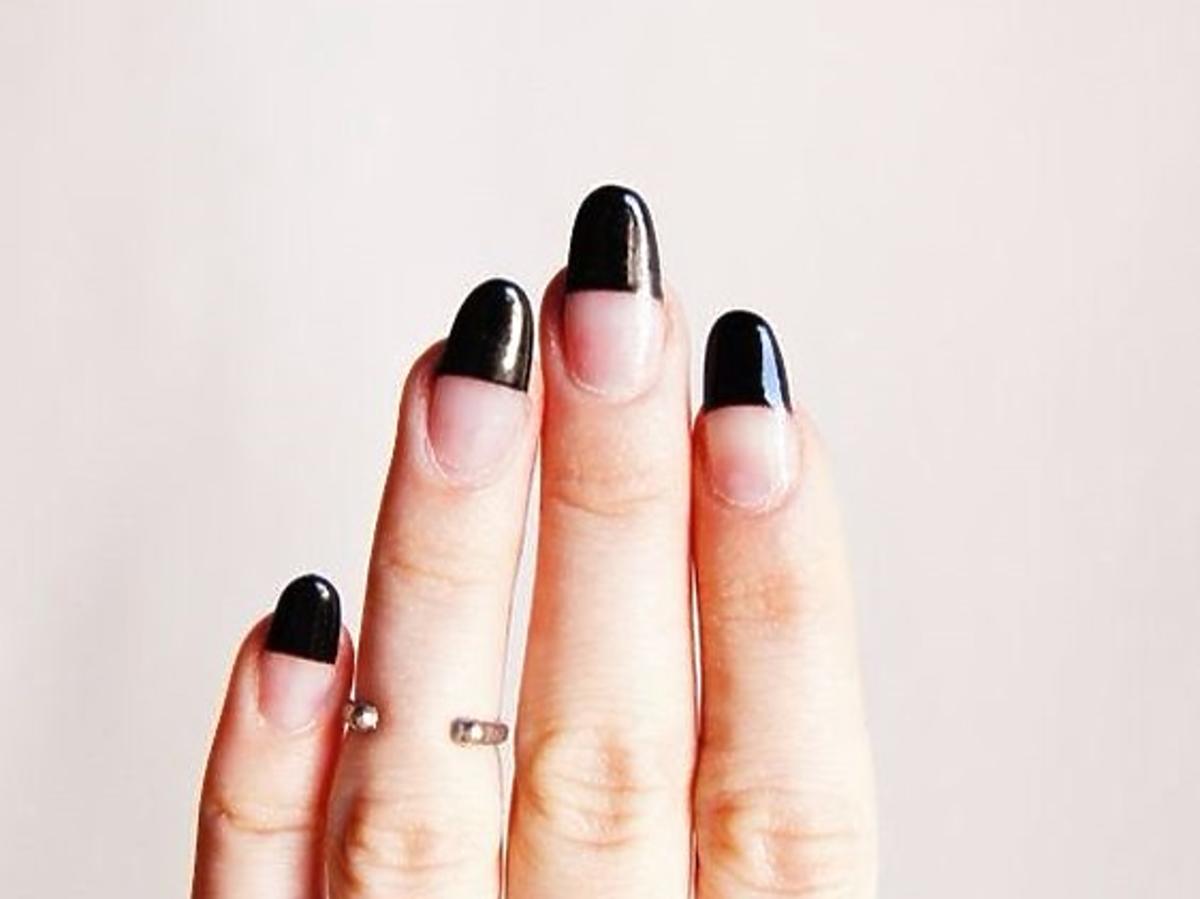 Dłoń na tle szarej ściany z paznokciami ze wzorem half-dip nails i złotymi pierścionkami