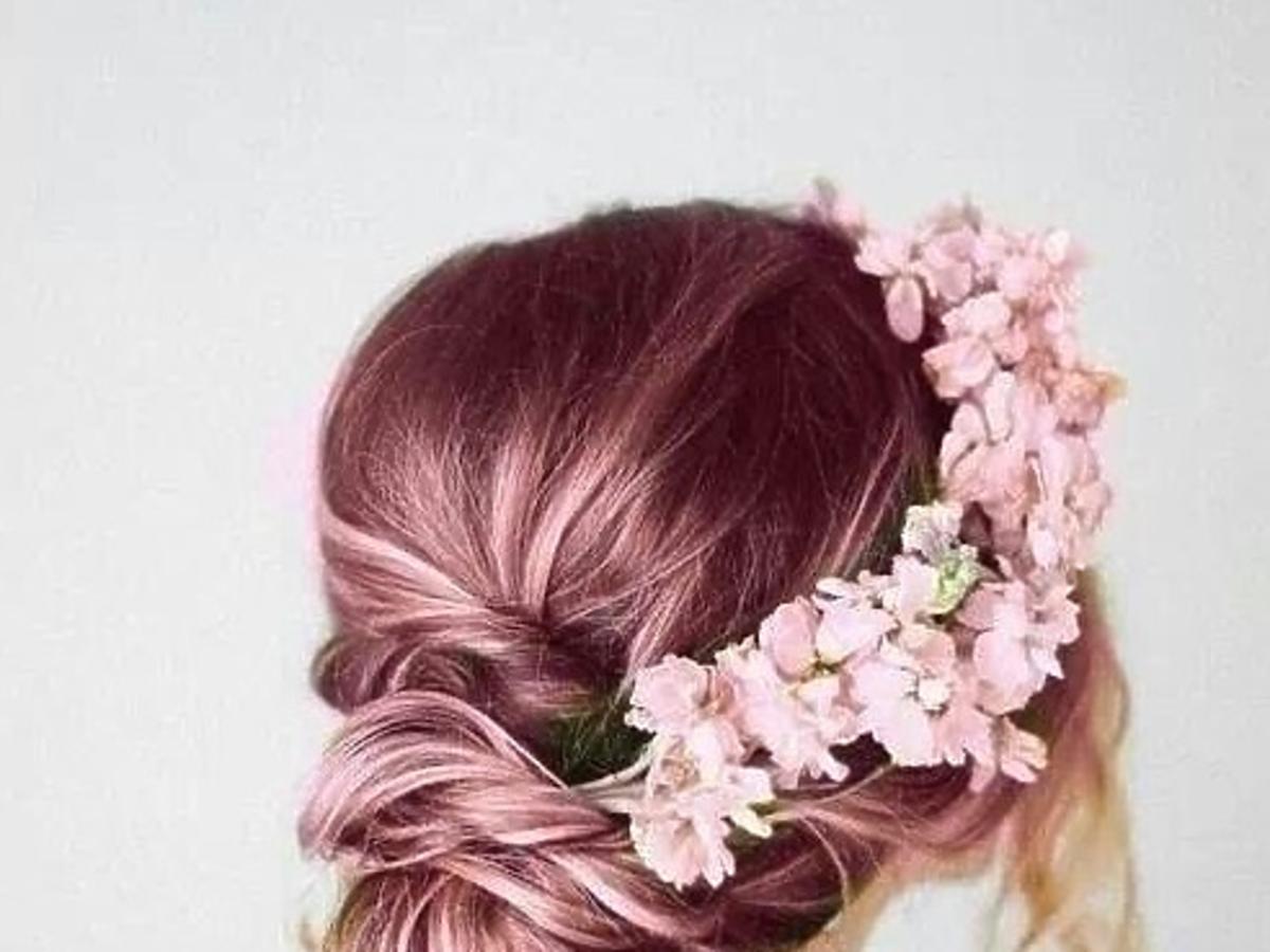 Dziewczyna z metaliczno różowymi włosami z wpiętym w nie wiankiem z kwiatów pozuje odwrócona tyłem na tle białej ściany