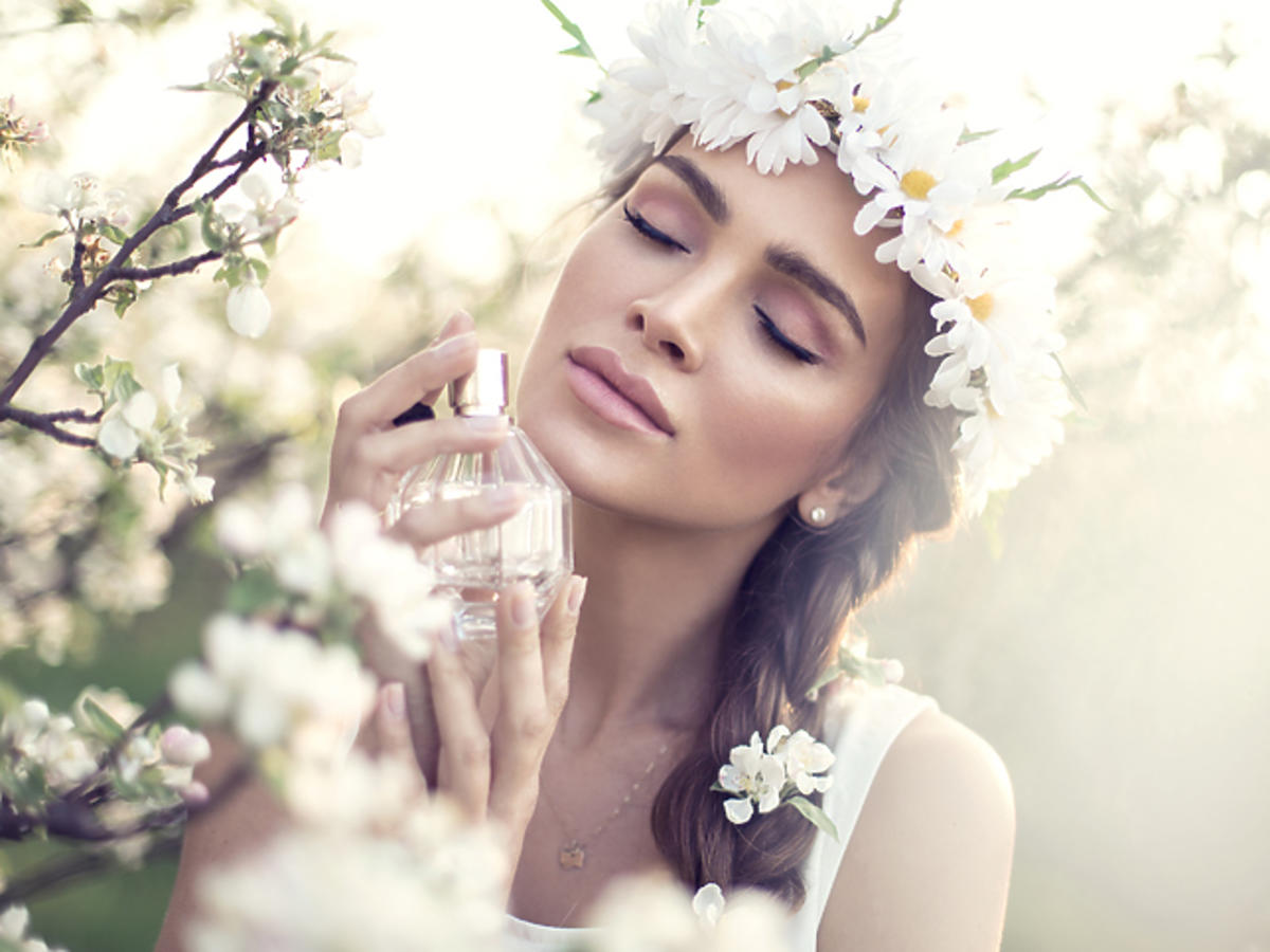 Kobieta z wiankiem w brązowych włosach na tle kwiatów wącha perfumy