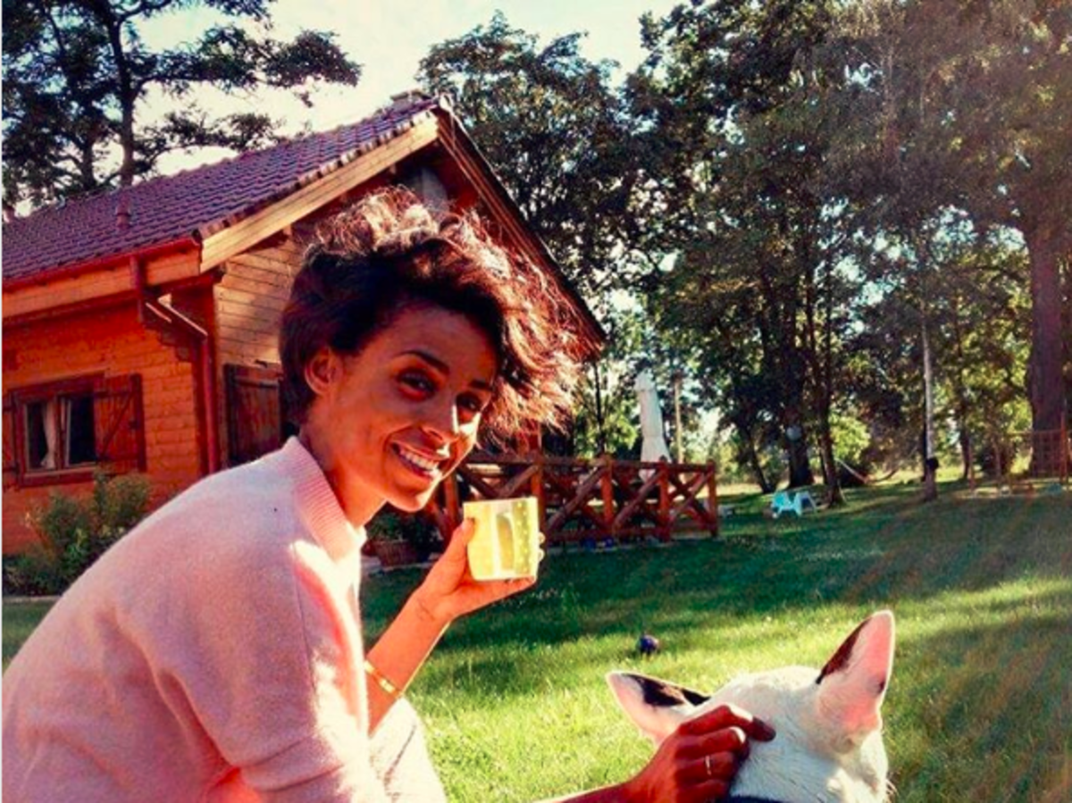Ola Szwed w długich włosach, ze szklanką w ręce, w różowym swetrze głask psa